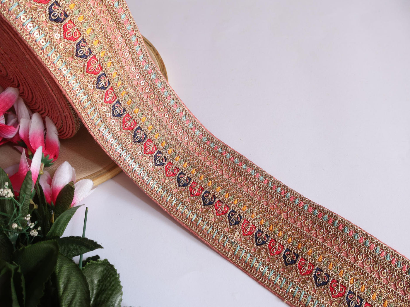 A yard of light peach zari sequin embroidered border trim with floral patterns, displayed against a white background with a partial view of a floral arrangement to the left.