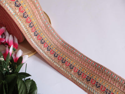 A yard of light peach zari sequin embroidered border trim with floral patterns, displayed against a white background with a partial view of a floral arrangement to the left.